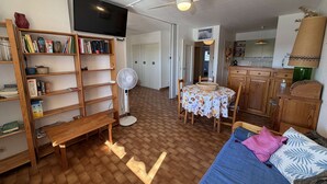 A cozy living room with a wooden bookshelf, a dining table with chairs, a blue sofa, and a ceiling fan. - Spacious 3-bedroom apt with terrace, pool and parking near the beach (Port Barcarès)
