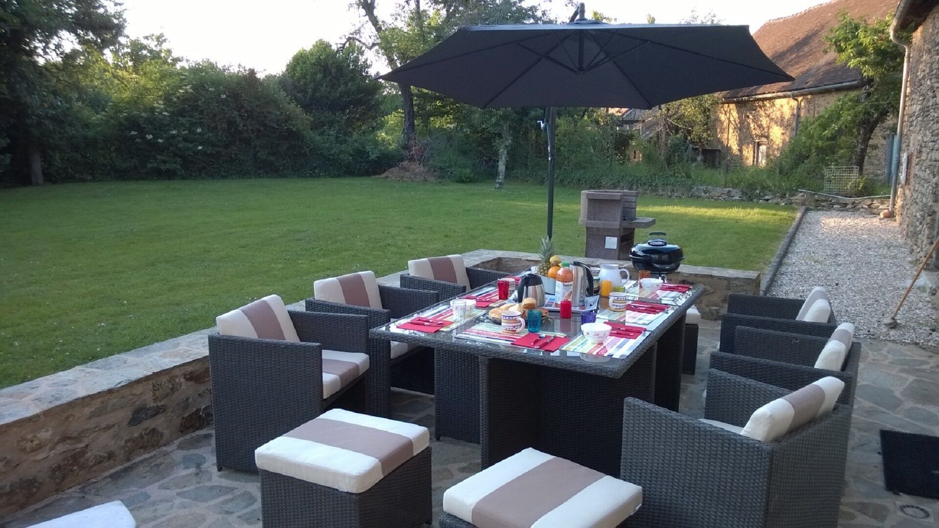 Um pátio externo com uma mesa posta para refeições, rodeado por cadeiras de vime, e um guarda-sol oferecendo sombra.
