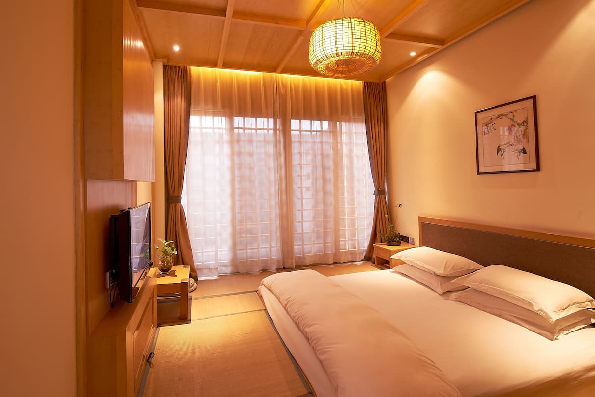 Ein Hotelzimmer mit einem großen Bett, einem Fernseher, einem Nachttisch mit Lampe und einem Vorhangfenster.