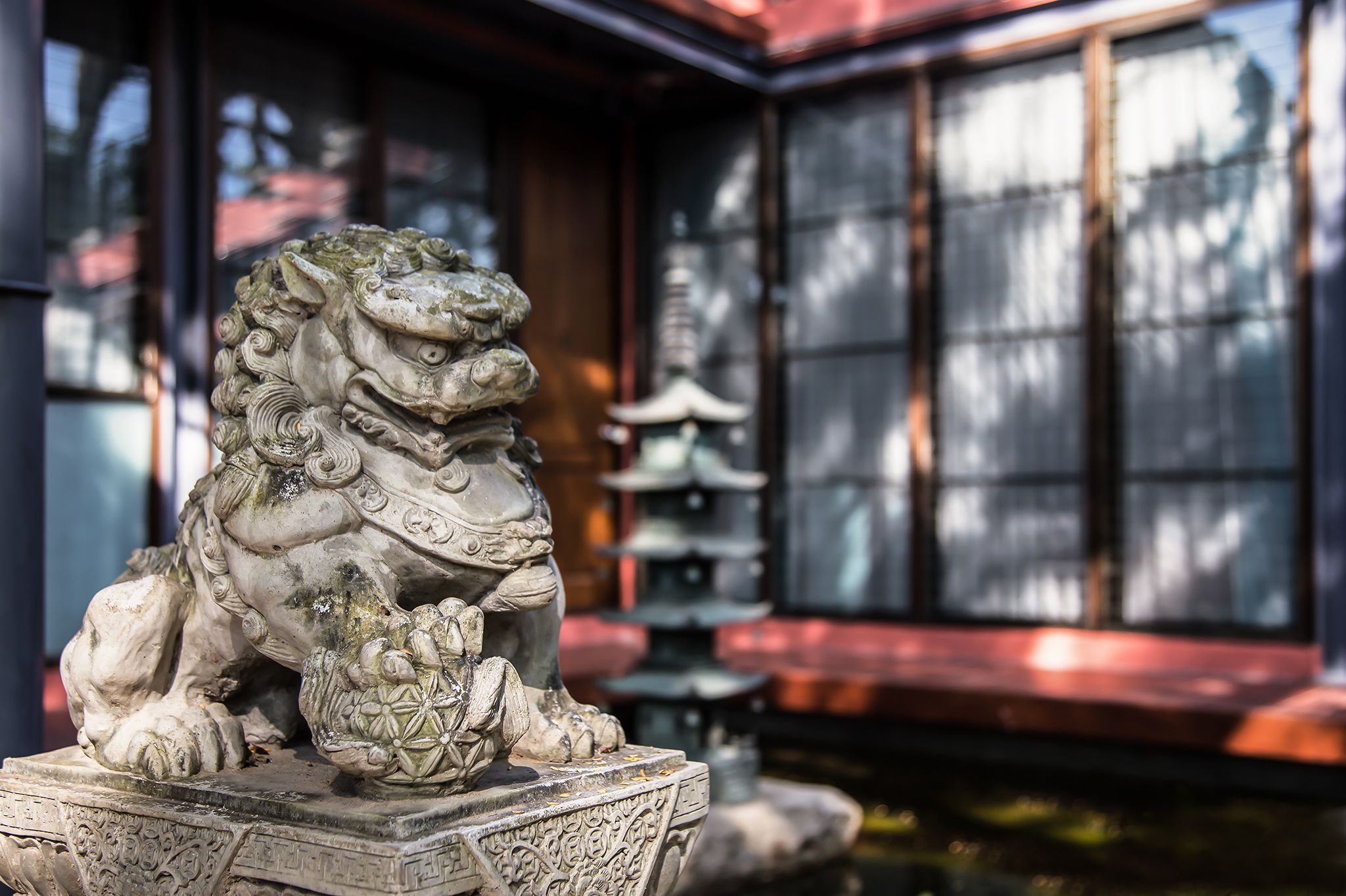 Una estatua de león de piedra en un patio con arquitectura japonesa tradicional, incluyendo una pagoda y una fuente.