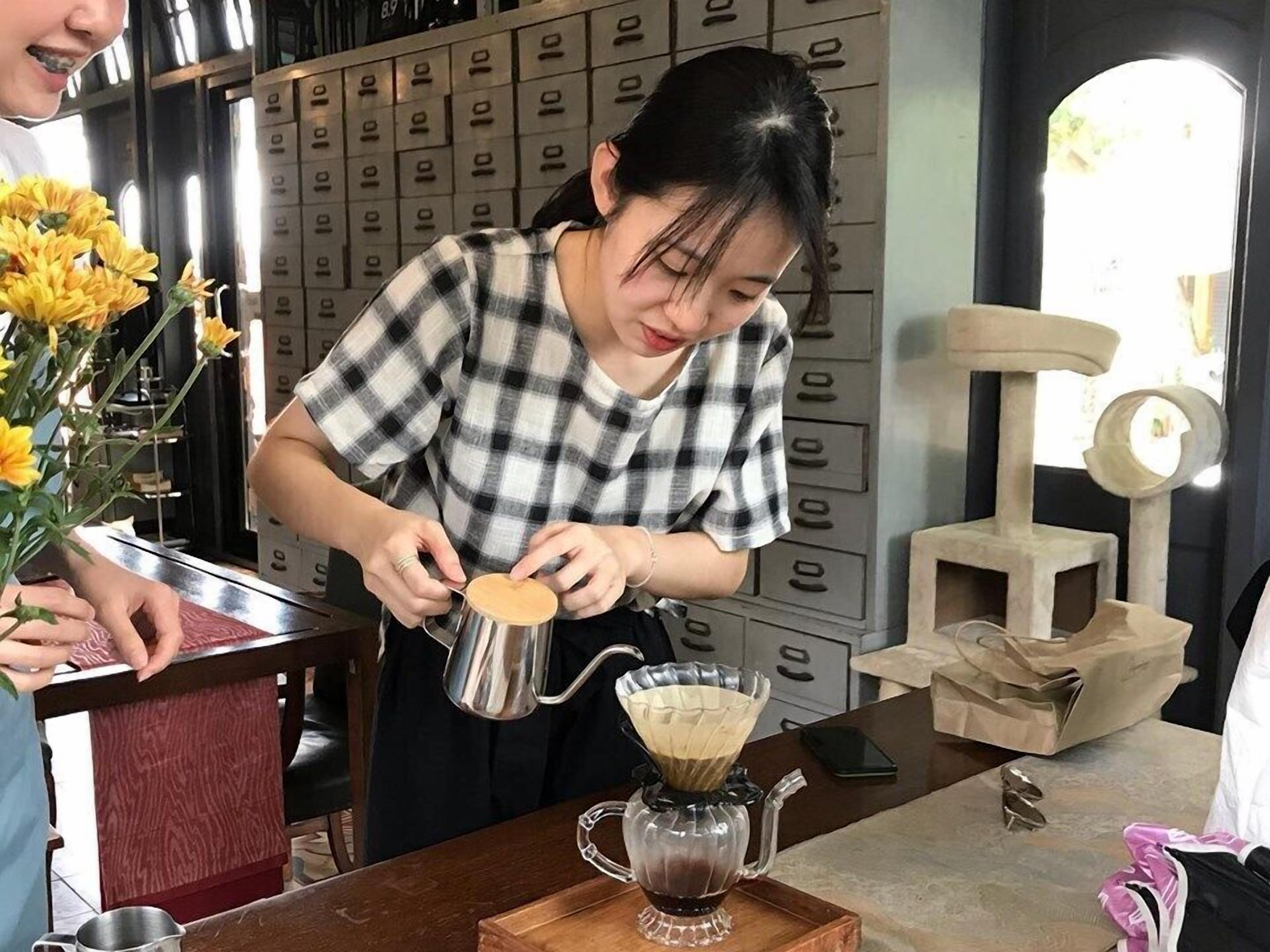 Una persona preparando café en una cocina con una cafetera, una mesa de madera y un rascador para gatos al fondo.