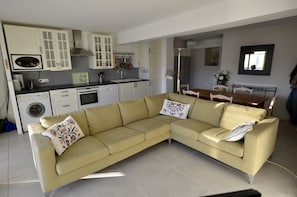Un salon moderne avec un canapé d’angle beige, un coin repas doté d’une table en bois et une cuisine avec des meubles et des appareils électroménagers blancs.