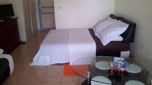 A hotel room with a bed, a glass table with a bottle and a brochure, a television on a wooden cabinet, and a door leading to another room. - Pretty studio in Sainte-Luce bordered by its beautiful beaches (Sainte-Luce)