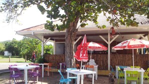 An outdoor seating area with tables and chairs under a tree, a covered area with a bamboo fence, and a building with a tiled roof in the background. - Pretty studio in Sainte-Luce bordered by its beautiful beaches (Sainte-Luce)