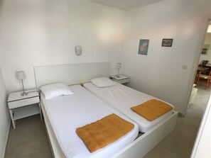 A double bed with white bedding and two brown throw blankets. There are two bedside tables with lamps, and a small wall-mounted light. The room has white walls and a tiled floor. - Cozy 2BR Villa in Port Leucate, 500m from Beach, Terrace, BBQ, Garage, Sleeps 5 (Port Leucate)