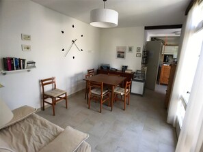A dining area with a wooden table and chairs, a white wall clock, and a kitchen in the background. - Cozy 2BR Villa in Port Leucate, 500m from Beach, Terrace, BBQ, Garage, Sleeps 5 (Port Leucate)
