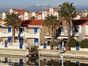 A coastal hotel with balconies, palm trees, and a mountain view. - Beautiful 4-person apartment with pool, private parking and marina view (Port Barcarès)