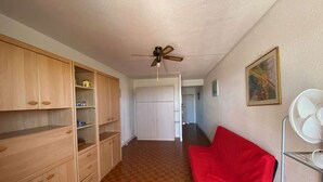 A room with a red sofa, wooden cabinets, a ceiling fan, and a tiled floor. - Seaside studio for 3 pers with parking (Port Leucate)