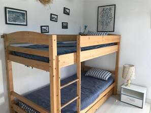 Uma cama de beliche com três andares, uma pequena mesa de cabeceira com abajur e várias fotos emolduradas na parede.