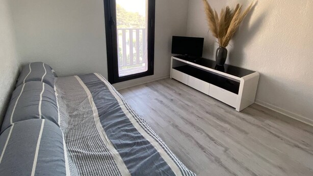 A bedroom with a bed, a TV on a white stand, and a window with a view of a balcony.