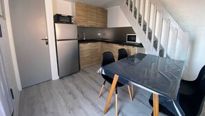 A compact kitchen with a refrigerator, microwave, and wooden cabinets. A dining table with chairs is positioned near a staircase.