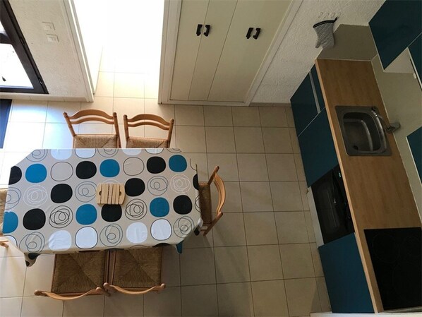 Zona pranzo con tavolo ricoperto da una tovaglia fantasia, sedie in legno e angolo cottura con pensili e lavello.