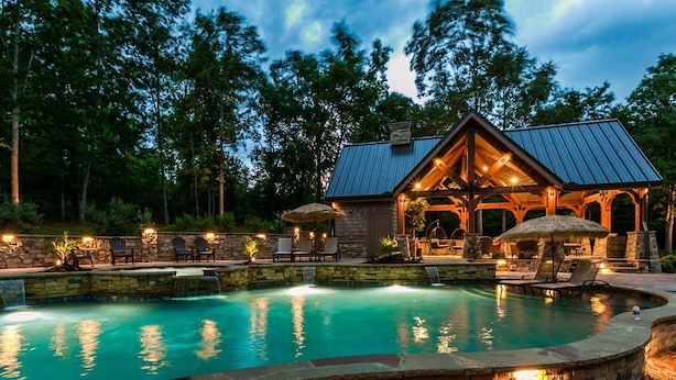 Area piscina con pareti in pietra e un gazebo, immersa tra alberi e un edificio con tetto in metallo.
