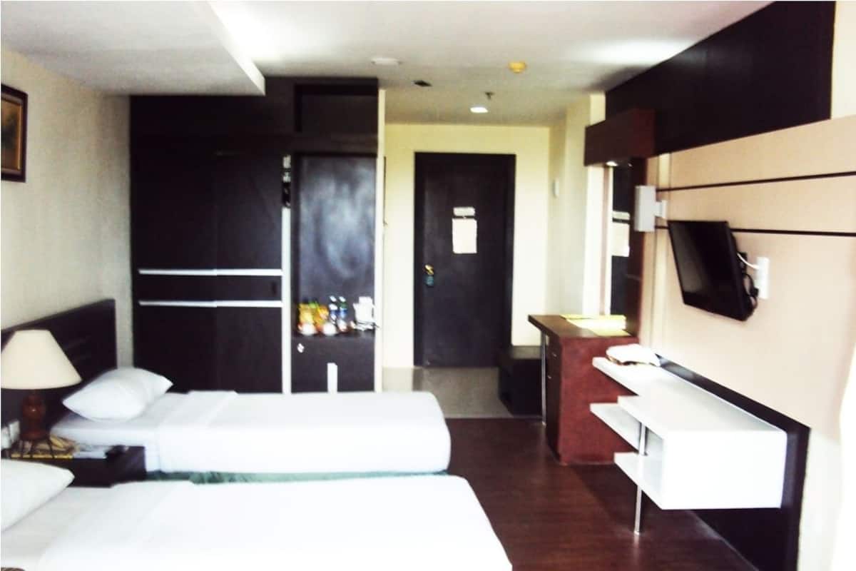 A hotel room with two beds, a desk, a TV, and a door leading to a hallway.