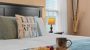 Ein ordentlich bezogenes Bett mit Kissen, ein Tablett mit Speisen und einer Tasse, eine Lampe und ein Fenster mit Jalousien.