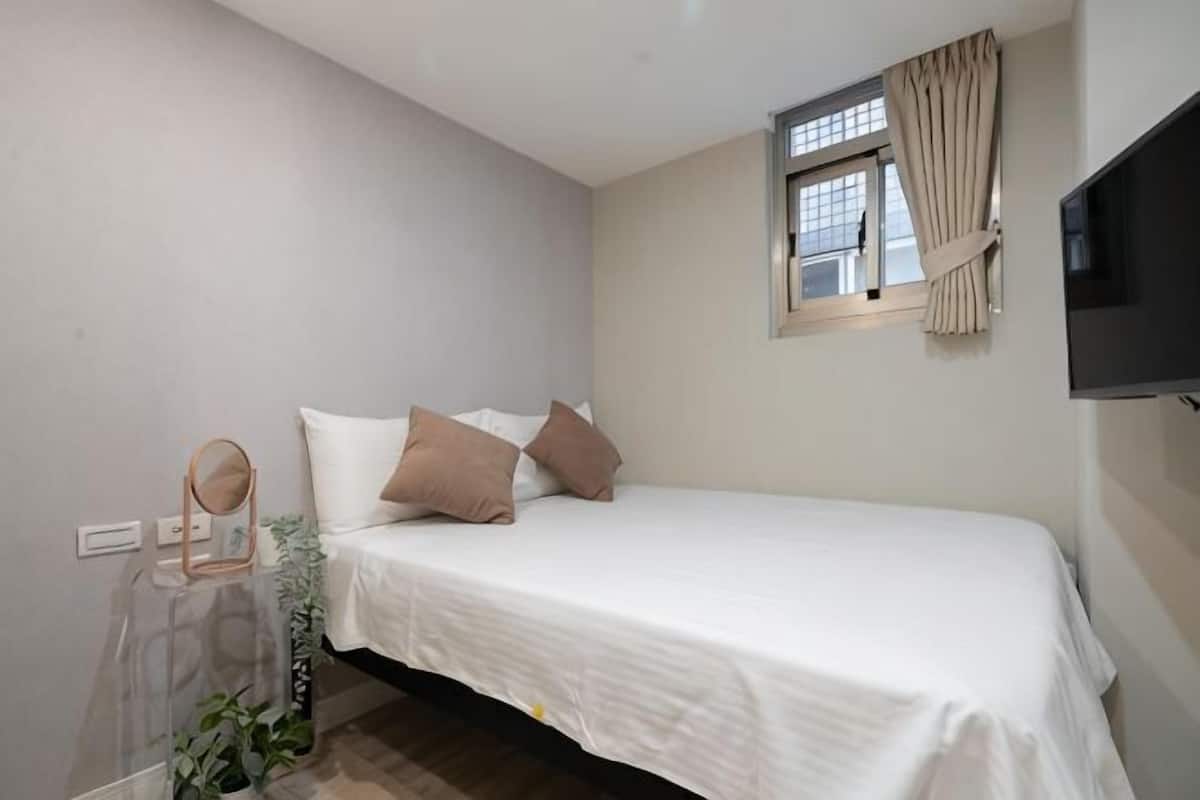 A bedroom with a bed, a TV, a window with curtains, and a bedside table with a mirror.