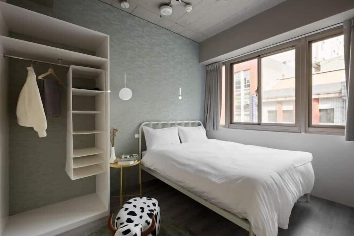 A bedroom with a bed, a wardrobe, a small table, and a window with a view of buildings.