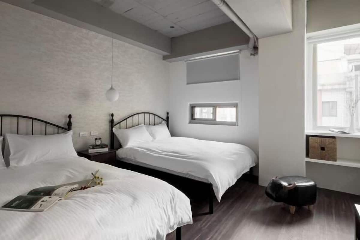 A modern bedroom with two beds, a small stool, a nightstand, and a window with a view of buildings.