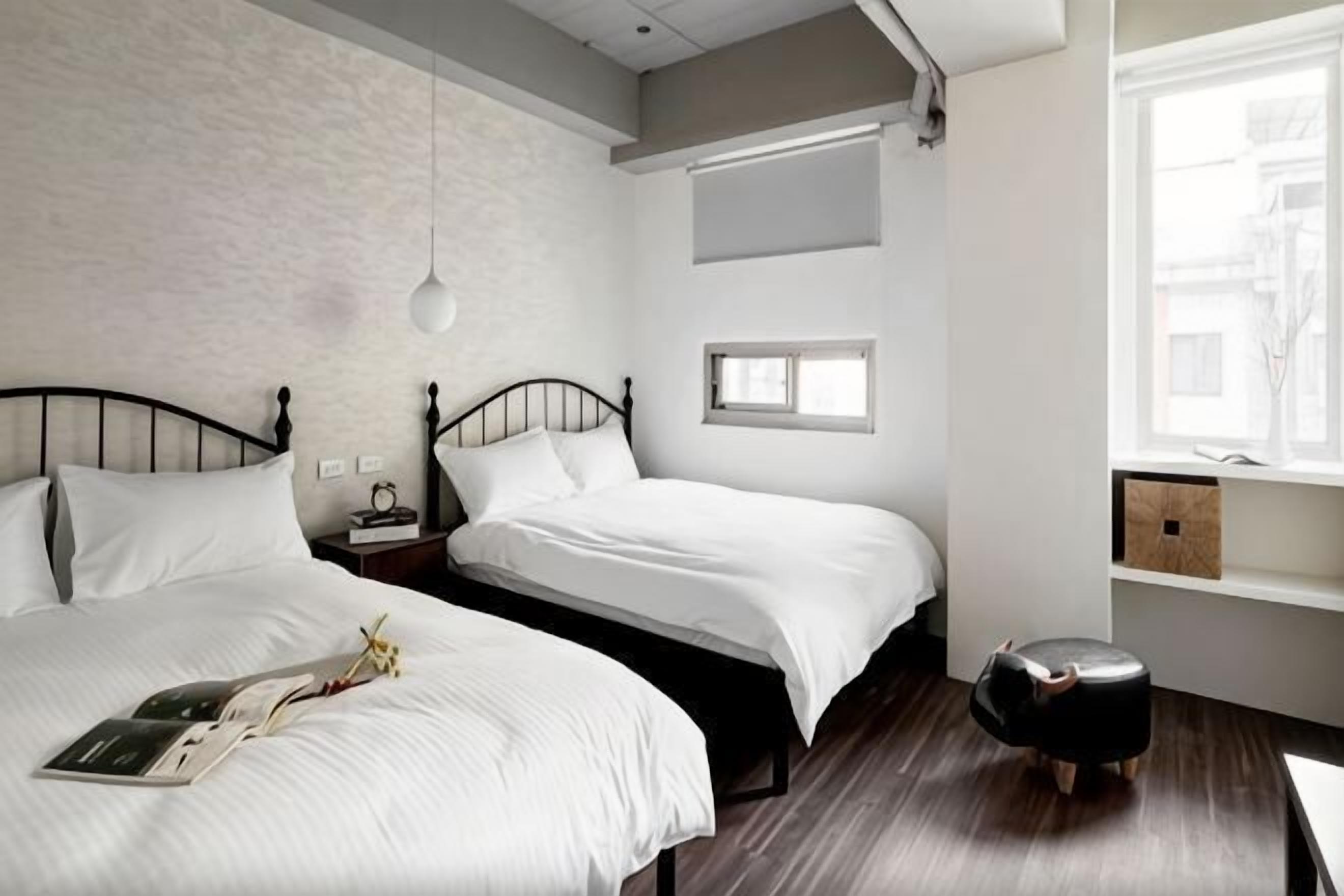 A bedroom with two beds, a bedside table with a lamp, a window, and a small stool.