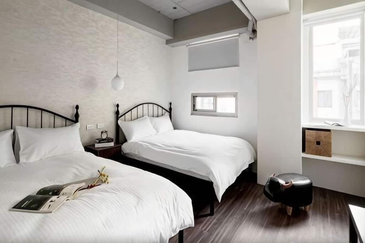 A bedroom with two beds, a bedside table with a lamp, a window, and a small stool.