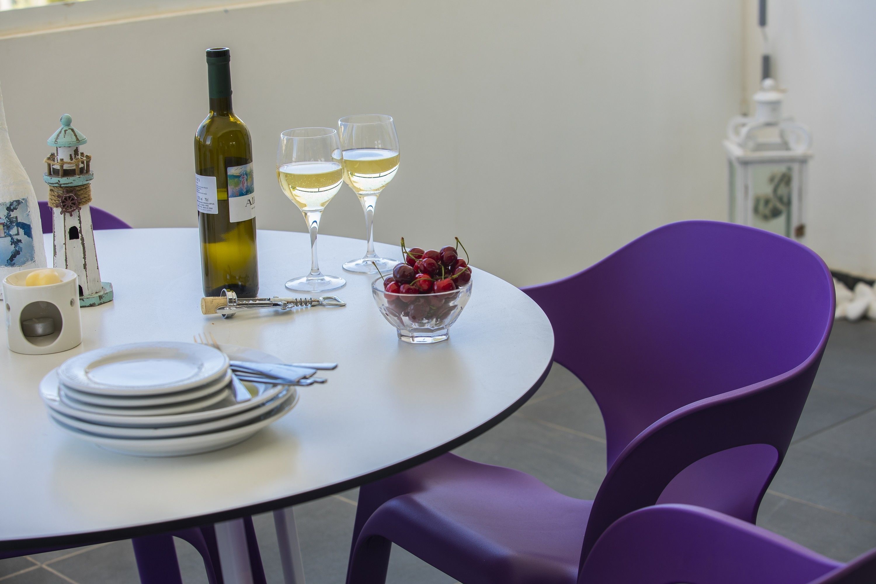 A round table with a white tablecloth, two wine glasses, a bottle of wine, a bowl of cherries, and a stack of white plates.