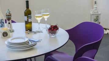Una mesa redonda cubierta con un mantel blanco, dos copas de vino, una botella de vino, un bol de cerezas y una pila de platos blancos.