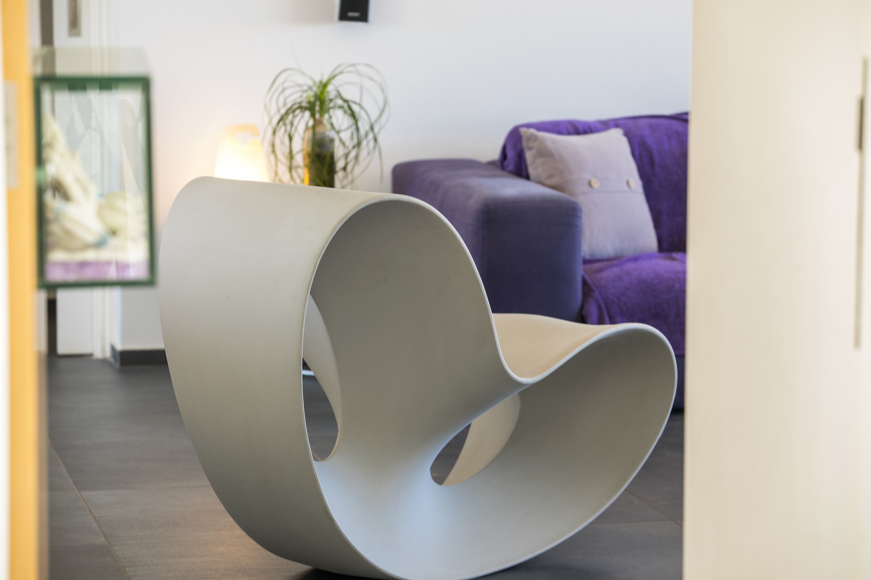 Una silla moderna y curvada en una habitación contemporánea con un sofá púrpura y un cuadro en la pared.