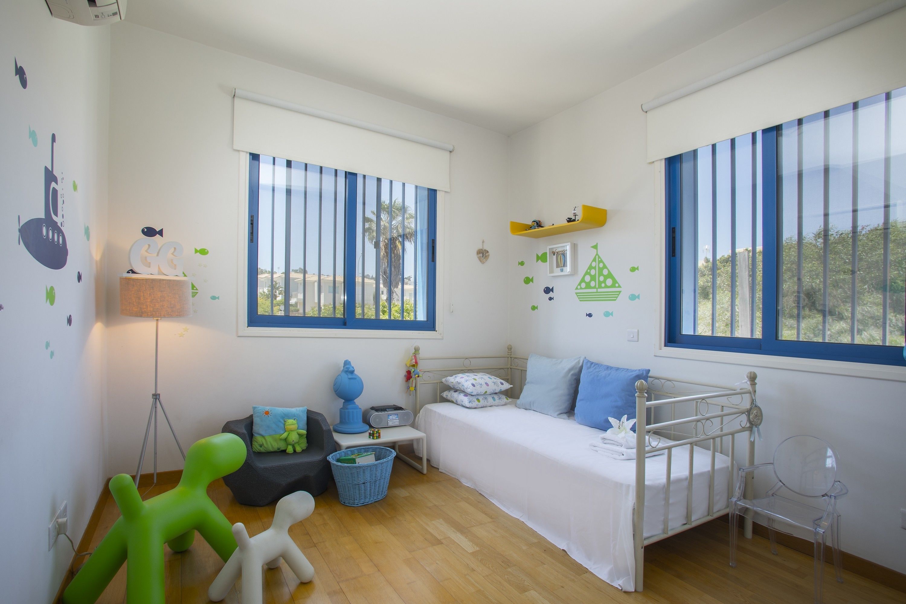 Habitación infantil con una cama, un escritorio, una lámpara y una ventana que da a vistas de palmeras.