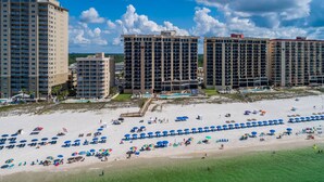 A beachfront hotel with multiple high-rise buildings, a sandy beach with numerous umbrellas, and a clear blue sky.