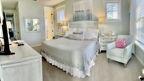 A bedroom with a large bed, bedside tables, a chair, and a dresser. There are two windows with blinds, a mirror, and a lamp on the bedside tables.