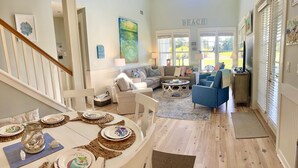 A spacious living room with a high ceiling, a staircase, a dining area with a table set for four, and a large window with a view of the outdoors.