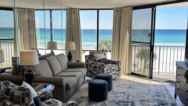 A spacious living room with a large window overlooking the beach, furnished with a sofa, armchairs, and ottomans.