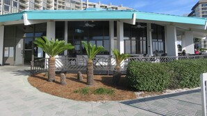 A modern building with a covered outdoor seating area, palm trees, and a paved walkway.