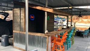 Ein Außenbereich mit Bar, einer Neonreklame, einem Grill und bunten Stühlen.