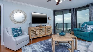 Ein Wohnzimmer mit grauem Sofa, blauem Sessel, Couchtisch aus Holz und einem Fernseher auf einer Holzkommode.
