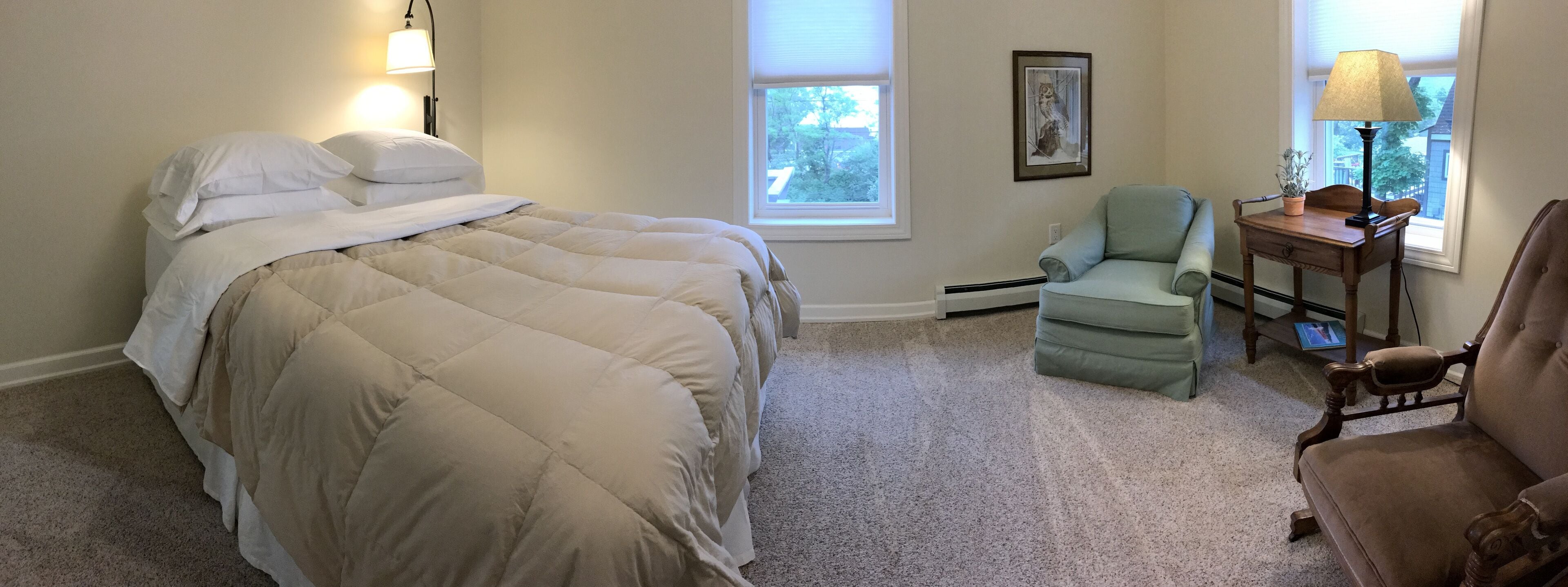 Un dormitorio con una cama grande, dos lámparas de noche, una silla, una mesita con lámpara y una ventana con persianas.