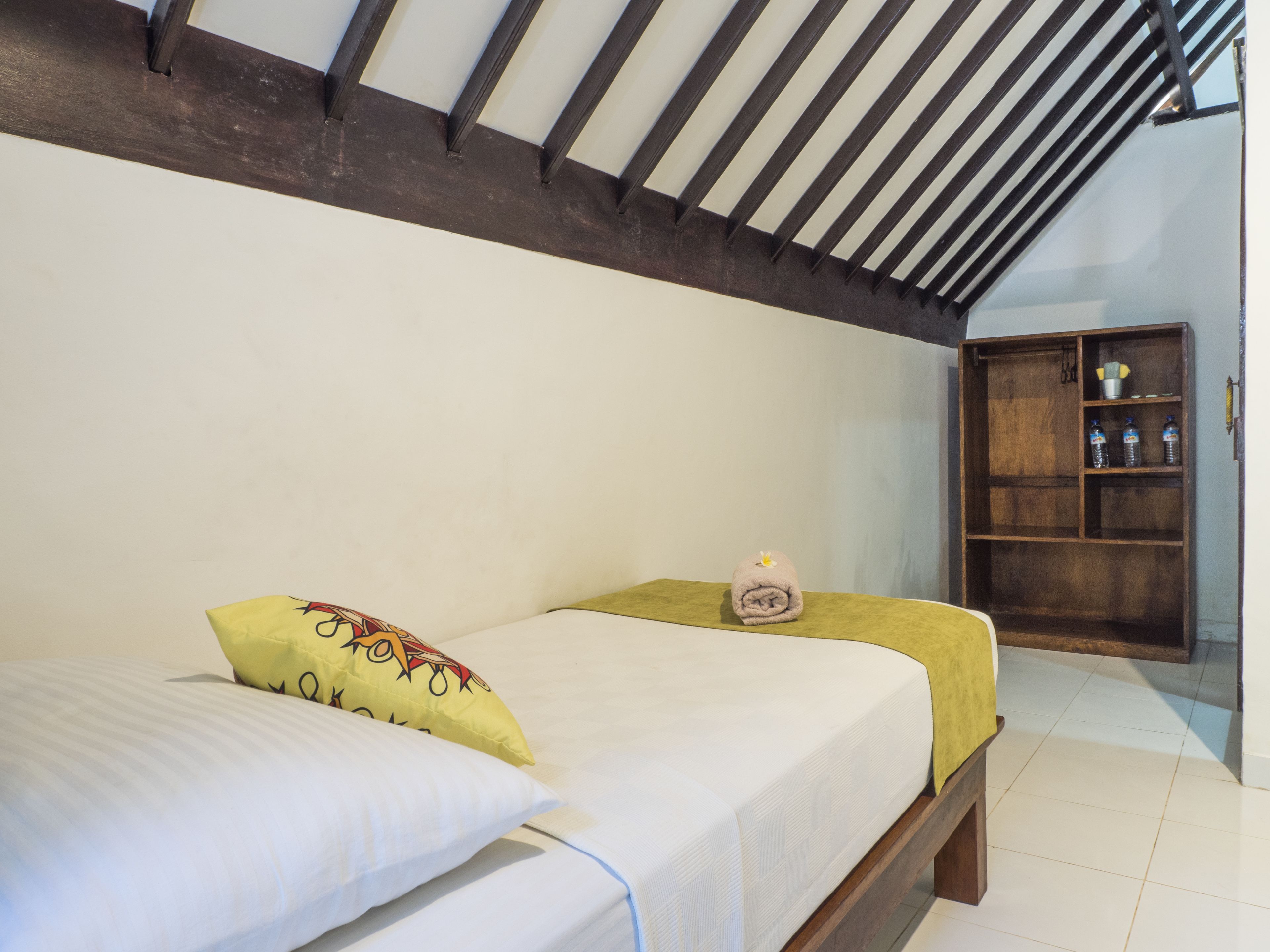 Une chambre à coucher avec un lit, des oreillers, une table de chevet, une étagère en bois avec des bouteilles et un plafond en pente.