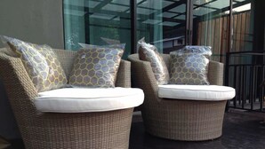 Dos sillones de mimbre con cojines blancos, sobre un piso de baldosas oscuras, con una puerta de cristal y una valla de madera al fondo.