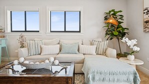 Un salon avec un canapé beige, deux fenêtres, une table basse agrémentée d’objets décoratifs, un lampadaire et une plante en pot.