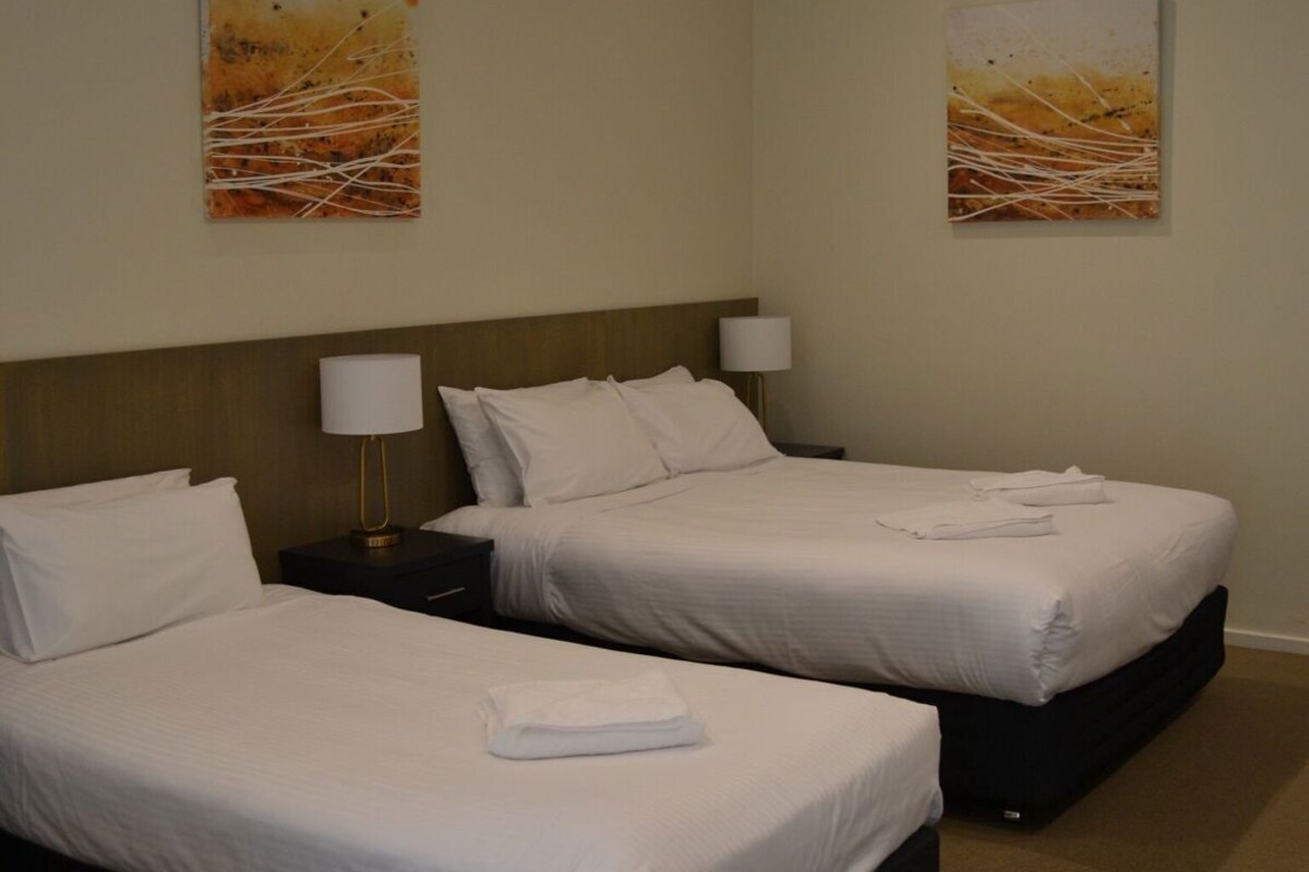 A hotel room with two beds, a headboard, bedside tables with lamps, and two wall-mounted paintings.