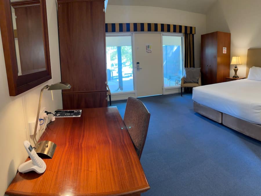 A hotel room with a bed, a desk with a telephone, a chair, a lamp, and a view of the outside.