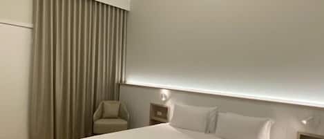 A modern hotel room with a large bed, a chair, a nightstand, and a built-in shelf.