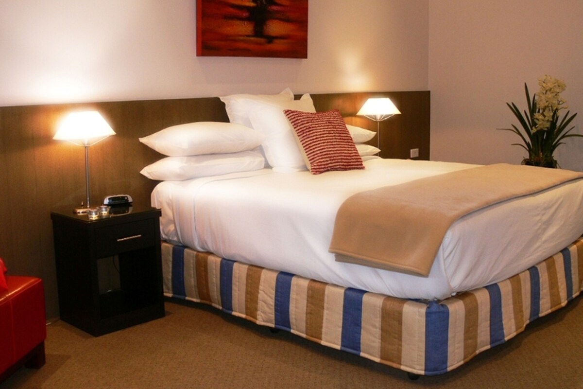A hotel room with a bed, two bedside lamps, a nightstand, a red chair, and a painting on the wall.
