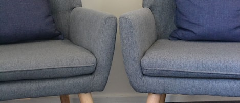 Two grey armchairs with wooden legs and blue cushions against a plain wall.