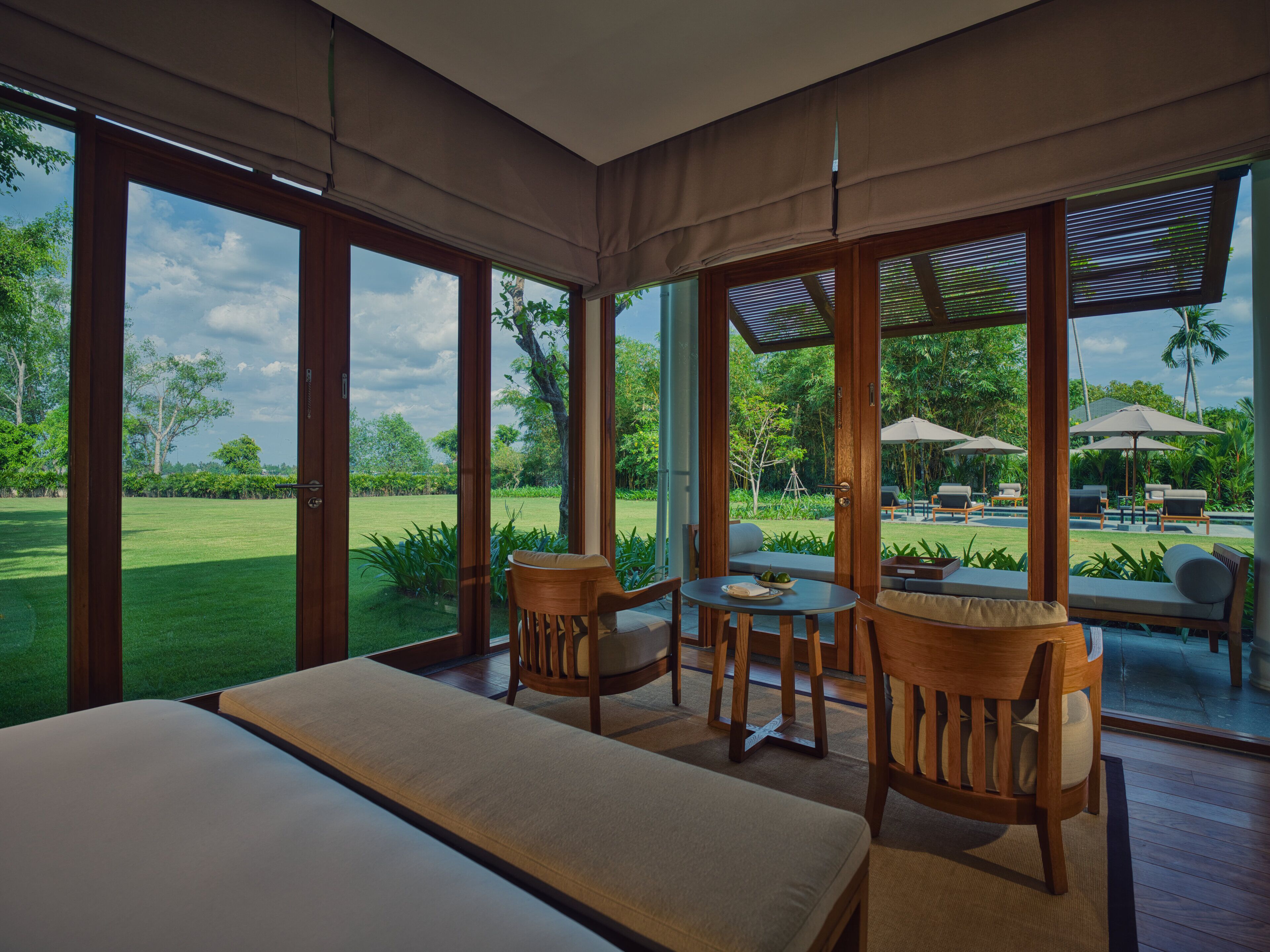 A spacious room with a large bed, two chairs, and a table. It features large windows offering a view of a lush garden and outdoor seating area.