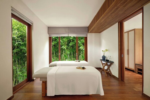 Une salle de spa équipée de deux tables de massage, d’un plancher en bois, de grandes fenêtres donnant sur un jardin de bambous et d’une petite table sur laquelle sont posés un vase et des bouteilles.