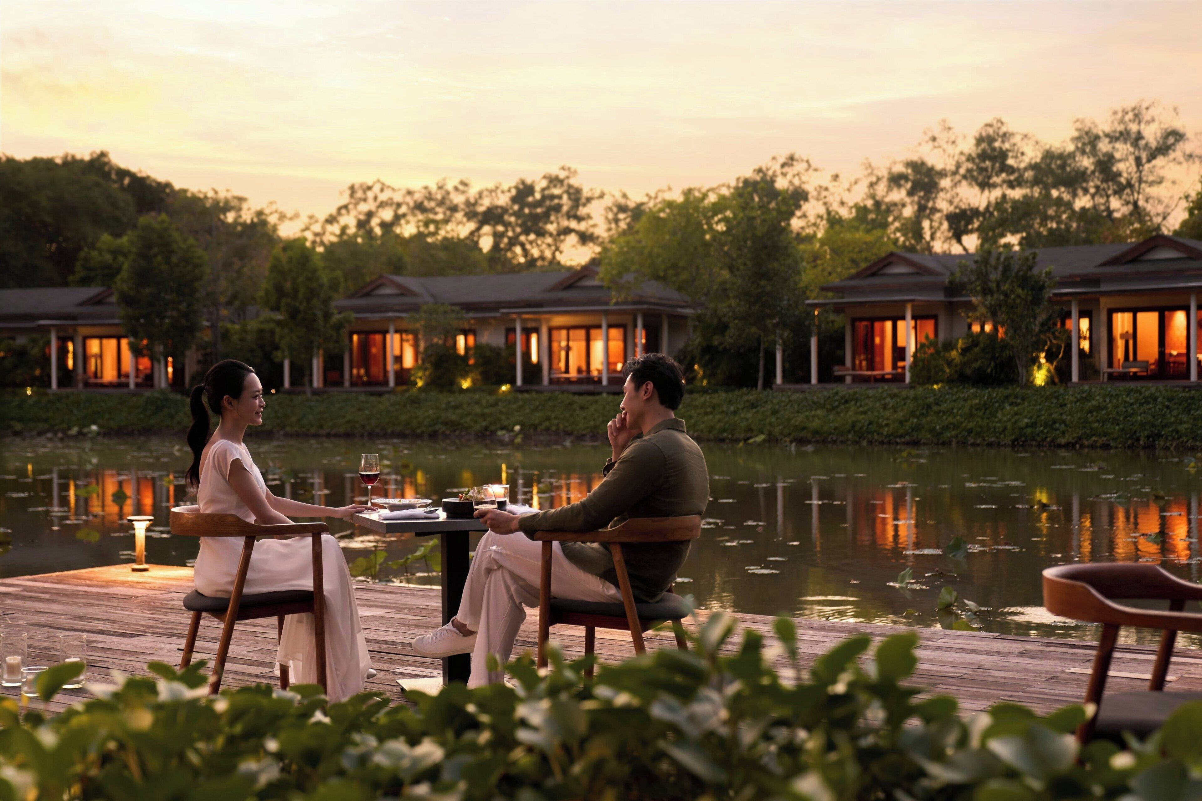Un couple est assis à une table sur une terrasse en bois donnant sur un lac, avec des bâtiments de style complexe hôtelier en arrière-plan.