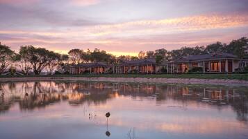 A row of waterfront villas with reflective water in the foreground, surrounded by trees and a sunset sky.