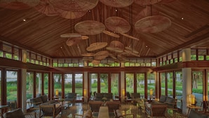 A spacious indoor area with multiple seating arrangements, large windows offering views of greenery, and a ceiling with hanging light fixtures. - Legacy Mekong, Can Tho, Autograph Collection (Can Tho)
