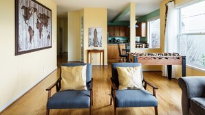 Sala de estar con dos sillones, una mesa de centro de madera y una mesa de futbolín. Se observa una cocina al fondo, con armarios de madera y un ventanal.